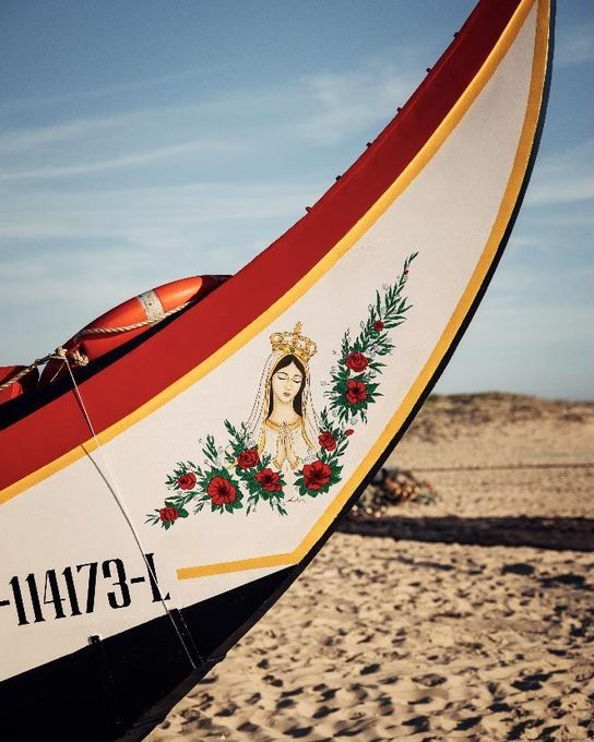 The prow of a traditional Aveiro boat on the sand: a white hull with red and gold trim, hand-painted with Our Lady of Fatima framed by red roses.