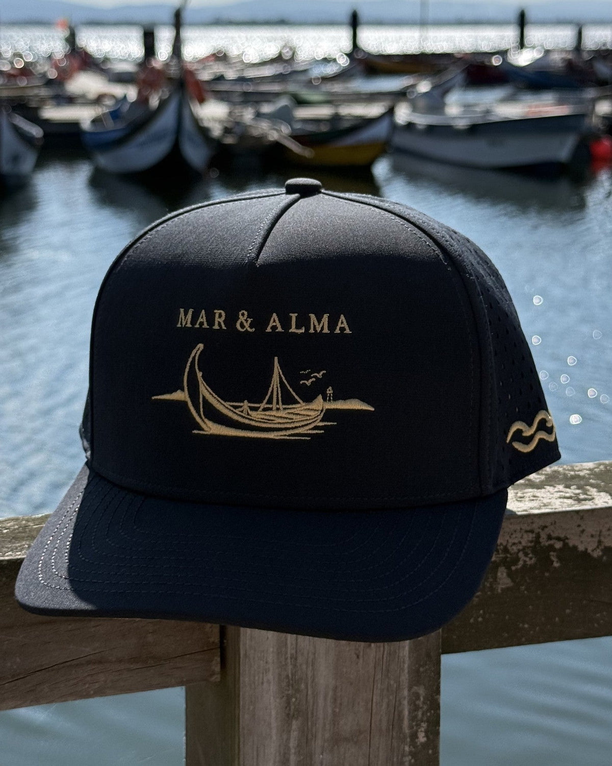 The Heritage Moliceiro Snapback in navy on a harbour rail, the moliceiro fleet moored behind.