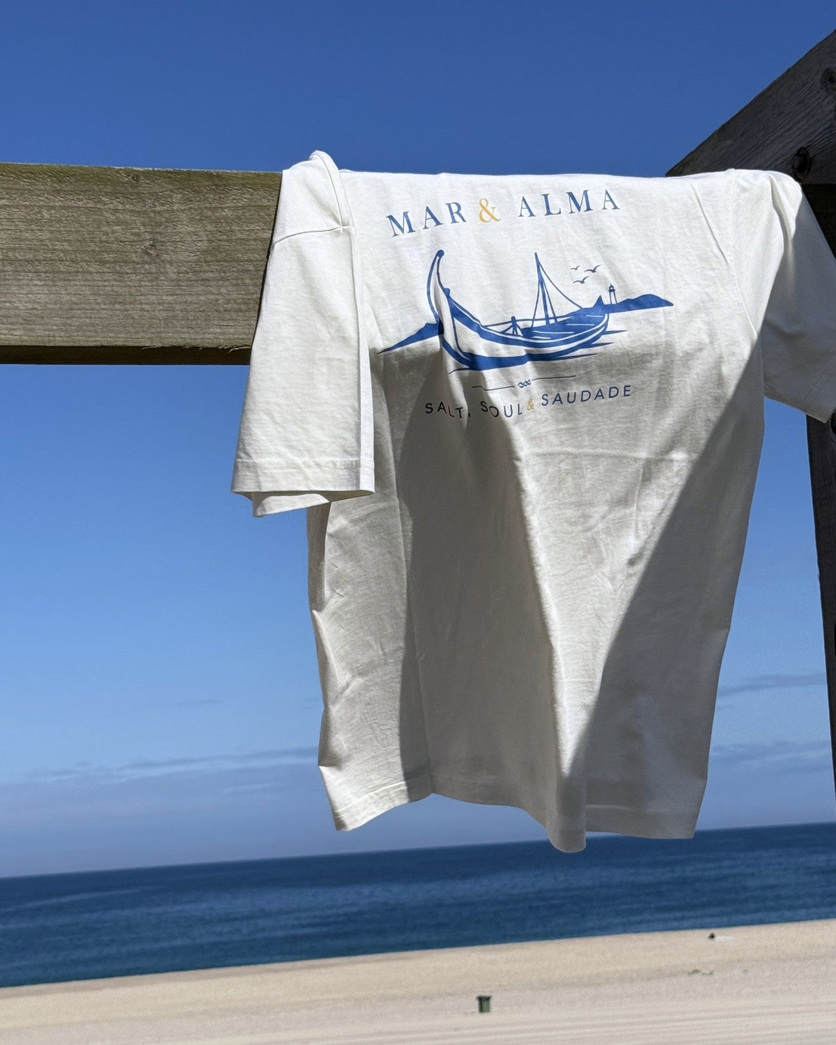 The Founders Tee hung on the boardwalk rail above the beach, the moliceiro print facing the sea.